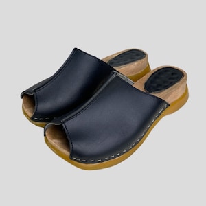 Pode incluir: Um par de tamancos pretos de couro com biqueira aberta. Os tamancos têm sola de madeira com fundo de borracha e costura branca na borda. Estes sapatos combinam conforto e estilo.