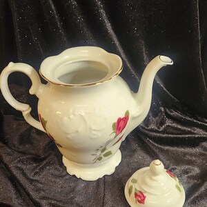 以下が含まれることがあります： 金色の縁取りとピンクのバラの花柄が施された白い磁器製のティーポット。ティーポットには、ノブ付きの蓋が付属しています。