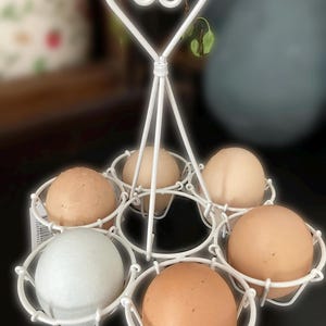 Könnte beinhalten: Weißer Eierhalter aus Metall mit herzförmigem Griff. Sechs Eier werden in dem Halter präsentiert, darunter braune, weiße und blaue Eier.