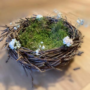 Könnte beinhalten: Ein rustikaler, kreisförmiger Kranz aus verflochtenen braunen Zweigen, gefüllt mit leuchtend grünem Moos. Zarte weiße Blüten und Zweige sind kunstvoll um den Kranz angeordnet und schaffen ein natürliches, dekoratives Stück. Ideal für die Inneneinrichtung.
