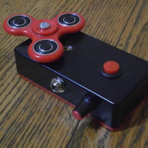 Könnte beinhalten: Ein schwarzes rechteckiges Gerät mit rotem Knopf, Drehknopf und Zierleiste sowie einem rot-schwarzen Fidget Spinner oben. Das Gerät hat einen silbernen Klinkeneingang und steht auf einer Holzoberfläche.