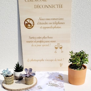 Peut inclure: Une pancarte en bois avec le texte "CÉRÉMONIE DÉCONNECTÉE" en français. La pancarte comprend également une icône interdisant les téléphones, un message demandant aux invités d'éteindre leurs téléphones et un message encourageant les invités à profiter de cette journée spéciale. La pancarte est posée sur une table avec une nappe blanche et quelques petites plantes en pot.