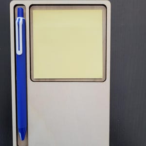 Può includere: Un organizer rettangolare in legno chiaro dotato di una fessura verticale sul lato sinistro contenente una penna a scatto blu con clip bianca, e un'area quadrata incassata sulla destra che ospita un blocco di foglietti adesivi giallo pallido.
