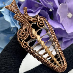 May include: A handcrafted copper wire pendant with a unique, openwork design. The pendant features a central, shell-like structure with intricate wire wrapping and decorative swirls. The piece is held against a backdrop of blue and purple flowers.