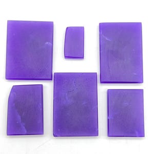Puede incluir: Colección de siete piezas de resina morada, rectangulares y con bordes curvos. Las piezas varían en tamaño y forma, con una apariencia suave y translúcida. La disposición sobre un fondo blanco resalta el color vibrante y la forma de la resina.