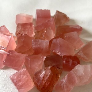 May include: A scattering of raw rose quartz crystals. The crystals are in various sizes and shapes, showing a spectrum of pink shades. The rough, unpolished surfaces reflect light, creating a shimmering effect. Suitable for jewellery making or crystal healing.