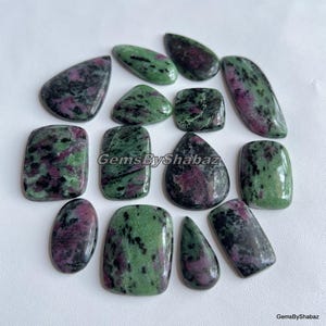 Könnte beinhalten: Eine Sammlung polierter Rubin-Zoisit-Edelsteine in verschiedenen Formen, darunter oval, rechteckig und tropfenförmig. Die Steine weisen eine Mischung aus grünen und dunkelrosa Farbtönen mit marmoriertem Aussehen auf. Der Text "GemsByShabaz" ist sichtbar.
