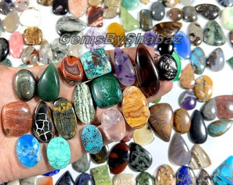 May include: A selection of polished gemstones in diverse shapes and hues, including green, blue, brown, and purple. The gemstones are held in a hand and scattered on a white surface. The text "GemsByShabaz" is visible.