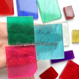 Könnte beinhalten: Eine Auswahl an farbenfrohen, durchscheinenden Edelsteinquadraten in Rot-, Blau-, Grün-, Rosa- und Lilatönen. Das Bild enthält den Text "GemsByShabaz". Diese polierten Steine werden wahrscheinlich für die Schmuckherstellung oder dekorative Zwecke verwendet.