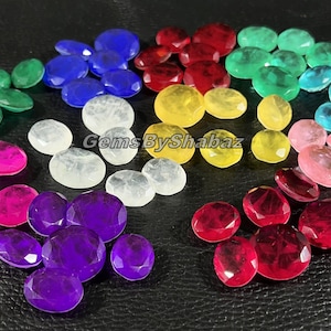 May include: An assortment of faceted gemstones in various colors, including green, blue, red, yellow, pink, and purple. The gemstones are arranged on a dark, textured surface, showcasing their polished surfaces and vibrant hues. The image is labeled "GemsByShabaz".