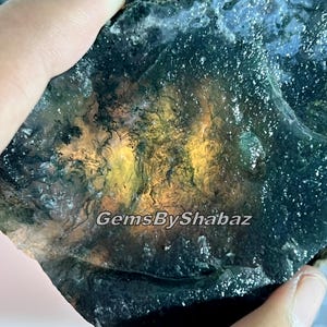 Puede incluir: Una gema en bruto con una mezcla de tonos verde oscuro, azul y amarillo dorado. La superficie tiene un aspecto texturizado, con áreas de translucidez. El texto "GemsByShabaz" es visible.