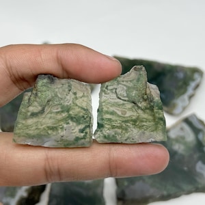 May include: Two pieces of raw moss agate gemstone, showcasing a blend of green and white patterns. The rough-cut stones have a natural, organic appearance, with visible textures and variations in color. Additional pieces are visible in the background.