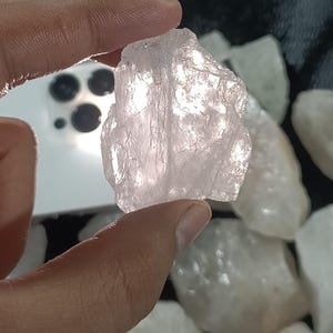 May include: A hand holding a rough, light pink rose quartz crystal. The crystal has a translucent appearance and a textured surface. Other similar crystals are visible in the background.