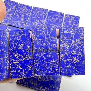 Może przedstawiać: Kolekcja prostokątnych, polerowanych kamieni szlachetnych. Kamienie mają żywy niebieski kolor podstawowy z uderzającym złotym wzorem żyłkowania. Tekst "GemsByShabaz" jest widoczny na kamieniach.