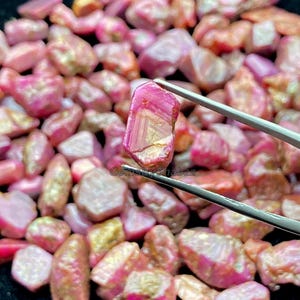 May include: A close-up of a collection of raw, uncut gemstones in shades of pink and gold. One gemstone is held by tweezers, showcasing its crystalline structure and colour variations. The background is filled with similar stones.