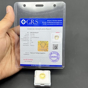 May include: A yellow cushion-cut sapphire gemstone, weighing 7.11 carats, is displayed in a clear plastic holder with a Gemstone Identification Report from GRS. The report details the gem's characteristics and origin.
