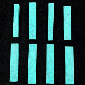 May include: Eight rectangular turquoise stones with a matrix pattern of grey veins. The stones are arranged on a black surface. These stones are likely for jewellery making or other craft projects.