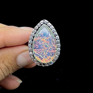 May include: A teardrop-shaped ring with a silver band and a large, iridescent gemstone. The gemstone displays a mix of orange, blue, and purple hues. The ring is held against a black background.