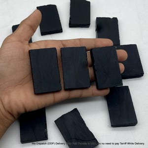 May include: A collection of small, rectangular black stone tiles with a smooth, matte finish. Several of these dark, uniform pieces are arranged in a scattered pattern, with three tiles resting directly on an open palm for scale.