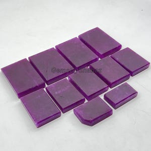 Puede incluir: Una colección de piedras preciosas rectangulares púrpuras pulidas. Las piedras están dispuestas en diagonal, con diferentes tamaños. La imagen está bien iluminada, mostrando el color y la textura de las piedras. El texto "GemsByShabaz" es visible.