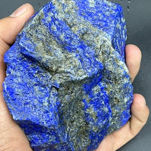 May include: A large, rough-hewn lapis lazuli stone, predominantly deep blue with streaks of gray and gold. The natural rock formation is held in a hand, showcasing its irregular shape and textured surface. A striking example of raw gemstone.