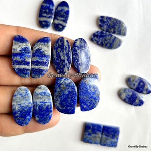 May include: A collection of polished lapis lazuli gemstones in various shapes, including ovals and rectangles. The stones display a deep blue color with streaks of gray and white, showcasing natural patterns. Some stones are held in a hand.