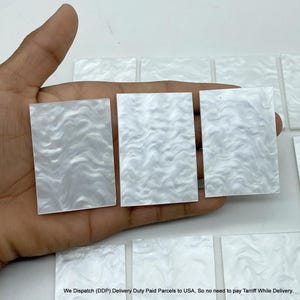 May include: A collection of rectangular white acrylic pieces featuring a swirling, pearlescent, marbled pattern. Each piece has a smooth, flat surface with a glossy finish, arranged in a grid-like formation on a plain white background.