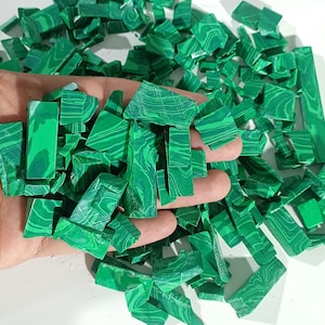 Pode incluir: Um punhado de peças de malaquite verde brilhante com veios verde escuro. As peças de malaquite são de várias formas e tamanhos, com uma superfície polida e lisa. As pedras preciosas são seguradas em uma mão, com mais peças espalhadas ao fundo.