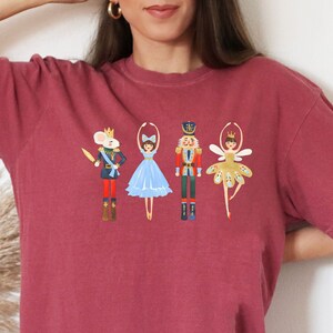 May include: Burgundy-colored t-shirt featuring a Nutcracker-themed design. The graphic includes illustrations of a mouse soldier, a ballerina in a blue dress, a nutcracker, and a fairy. The design is in a whimsical, cartoon style.