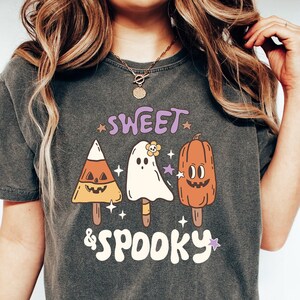 May include: Dark gray t-shirt with a Halloween-themed graphic. The design features candy corn, a ghost, and a pumpkin, all styled as popsicles, with the words "SWEET & SPOOKY" in a retro font. The shirt is worn by a person.