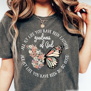 Pu&ograve; includere: Una maglietta grigia con un design a farfalla e fiori. Il testo sulla maglietta recita "All my life you have been faithful, goodness of God, all my life you have been so so good."