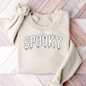 May include: A beige sweatshirt with the word "SPOOKY" in black, outlined letters. The letters have a dripping effect at the bottom. The sweatshirt has a crew neck and long sleeves with ribbed cuffs.