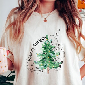 May include: An off-white short-sleeve t-shirt featuring a watercolor-style green Christmas tree graphic with colorful ornaments and the text Merry & Bright in black script, accented by black swirling lines and small dots.