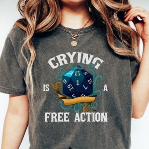 May include: A dark grey t-shirt with the text "Crying is a free action" printed on it. The text is in white and is surrounded by a blue and gold illustration of a 20-sided die with tentacles.