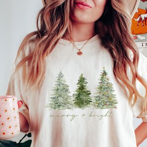 May include: Cream-colored t-shirt featuring a watercolor design of three Christmas trees. The trees are in shades of green, with the rightmost tree decorated with small ornaments. The words "merry + bright" are written in a script font below the trees.