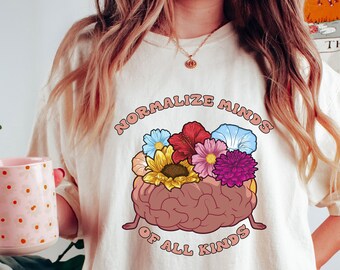 May include: Cream-colored t-shirt with the text "Normalize Minds of All Kinds" in a retro font. The graphic features a brain with colorful flowers. The person is holding a pink mug with white polka dots.