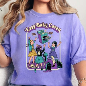 May include: Violet-colored t-shirt featuring the text "Easy-Bake Coven" with a graphic of three cartoon witches baking. The graphic includes a tombstone, a black cat, and an oven with baked goods. The shirt is a comfortable, casual style.
