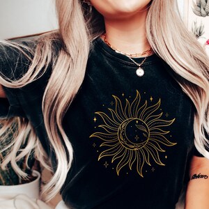 May include: A black t-shirt with a gold sun and moon graphic. The sun has rays and the moon is crescent shaped. The graphic is surrounded by small stars.