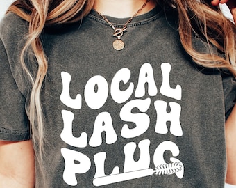 May include: A dark gray t-shirt with the text "LOCAL LASH PLUG" in white, stylized lettering. The text is in a retro font and includes a white mascara wand graphic.