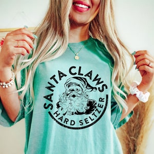 May include: A turquoise t-shirt with a black graphic that reads "SANTA CLAWS HARD SELTZER" in a circular design. The graphic features a Santa Claus illustration. The shirt has short sleeves and a relaxed fit.