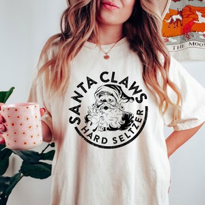 May include: Cream-colored t-shirt featuring a circular graphic with the text "SANTA CLAUS HARD SELTZER" and an illustration of Santa Claus. The shirt is a relaxed fit. A person is holding a pink mug with white polka dots.