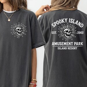 May include: Dark gray t-shirt with a white graphic of a skull inside a sunburst design on the front and back. The back of the shirt features the text "Spooky Island Amusement Park Island Resort" and the year "2002".