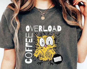 以下が含まれることがあります： チャコールグレーの半袖Tシャツで、大きな目をした黄色いフクロウのグラフィックが描かれています。テキストは白と黒の文字で「COFFEE OVERLOAD」および「MORE!」と書かれています。デザインは太い黒の輪郭線を用いた手描きのスケッチ風の仕上がりです。