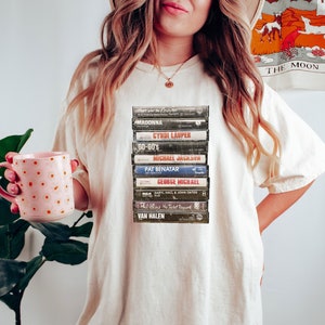 May include: A cream-coloured t-shirt displaying a stack of vintage cassette tapes, including artists such as Madonna and Michael Jackson. The tee has a relaxed fit and a crew neck. The design has a retro music theme, evoking nostalgia for the 80s and 90s.