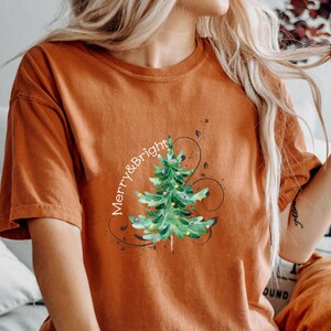 May include: A burnt orange short-sleeve t-shirt featuring a watercolor-style green Christmas tree graphic with colorful ornaments and the text Merry & Bright written in white script, accented by black swirling line art designs.