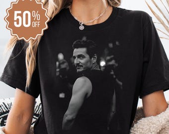May include: Black t-shirt featuring a black and white image of a man looking over his shoulder. The shirt has a round neckline and short sleeves. A brown, scalloped-edged sticker with the text "50% OFF" is in the upper left corner.
