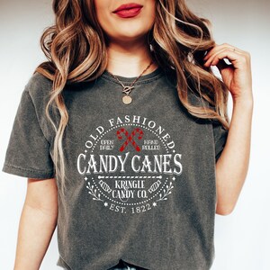 Pu&ograve; includere: T-shirt grigio scuro con grafica circolare che recita "Old Fashioned Candy Canes" con illustrazioni di bastoncini di zucchero rossi. Il testo include anche "Kringle Candy Co. Est. 1822". La maglietta &egrave; di vestibilit&agrave; ampia.