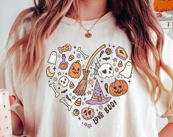 May include: Cream-colored t-shirt featuring a heart-shaped Halloween design with pumpkins, ghosts, a witch's hat, and the text "Love Boo!". The shirt is made of a soft material and has short sleeves.