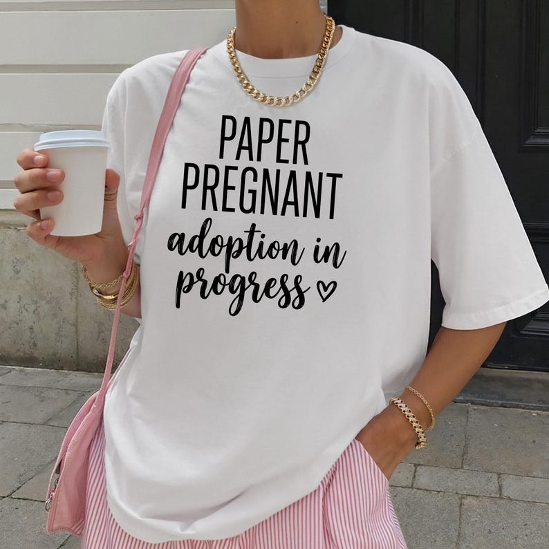 Pode incluir: Camiseta branca com o texto "PAPER PREGNANT adoption in progress" em preto. A camisa &eacute; combinada com cal&ccedil;as listradas rosa e branco e uma bolsa rosa. Um colar de corrente de ouro e pulseiras tamb&eacute;m s&atilde;o vis&iacute;veis.