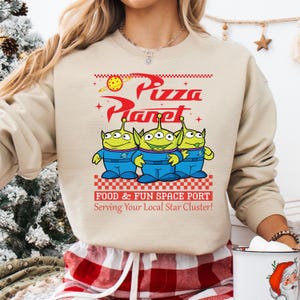 May include: A beige sweatshirt with a graphic of the Pizza Planet restaurant from the Toy Story movie. The graphic features the restaurant's logo, three alien toys, and the text "Food & Fun Space Port Serving Your Local Star Cluster!"